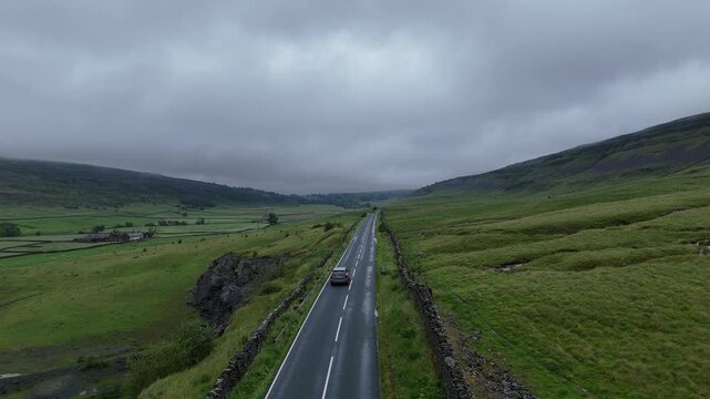 Aerial view of a car travelling on a wet road through lush green fields under a cloudy sky, Hawes, England, United Kingdom.