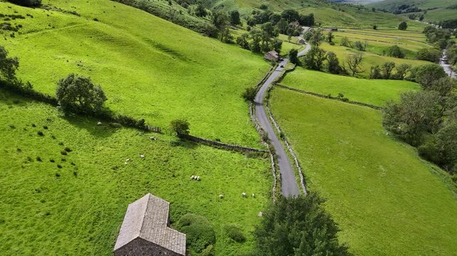 Aerial view of a stone barn nestled amid vibrant green fields, alongside a winding road that cuts through the landscape, Hawes, England, United Kingdom.