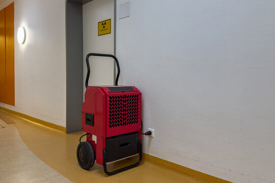 A red dehumidifier sits against a wall in a hallway, with a sign indicating a restroom nearby. The space features smooth flooring and clean, simple lines, creating a functional atmosphere.