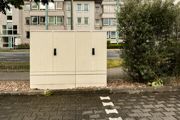 Utility boxes are positioned along a paved path next to modern residential buildings. Greenery...