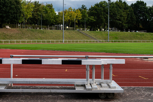 A track field features hurdles placed on the red running surface. In the background, lush green grass is visible, surrounded by trees and sports facilities. The scene is calm and clear.