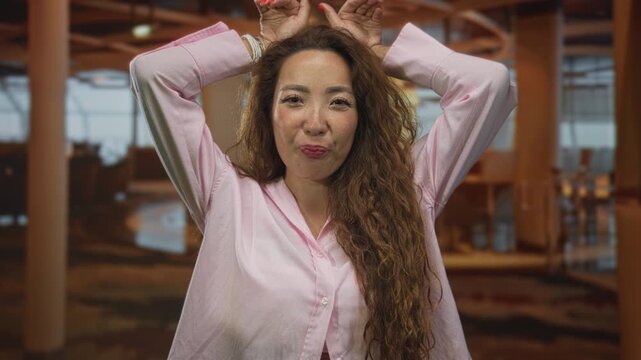 Woman with hands above head making a playful ear gesture and puckered lips in building; playful spontaneity.
