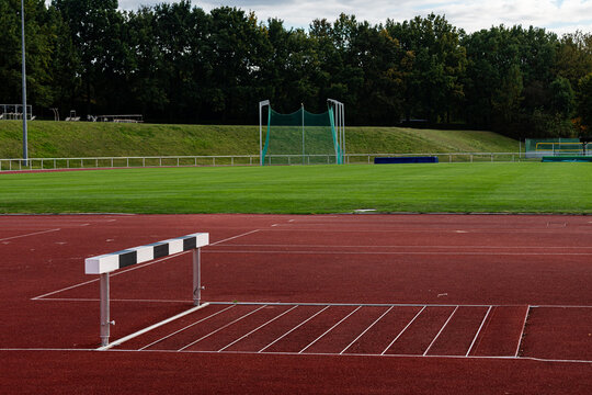 A clear view of a hurdle positioned on a red track at an athletic facility. The green grass field features trees in the background, providing a peaceful training atmosphere.