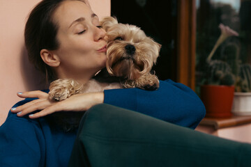 Woman kissing her adorable Yorkshire Terrier dog indoors. A woman is embracing and kissing her small Yorkshire Terrier dog, showing affection. The dog is looking at the camera with a cute expression.