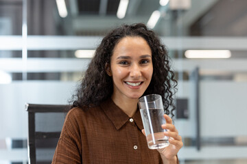 Young professional woman with curly hair smiling at the camera while drinking a glass of pure water...