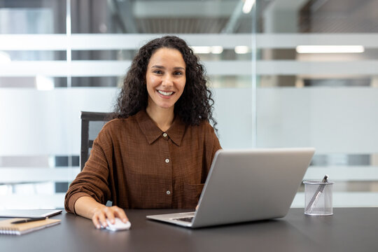 Young businesswoman with curly hair smiling at camera, using a laptop and mouse while sitting at a dark desk in a contemporary corporate office environment - Powered by Adobe