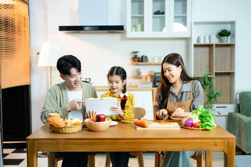 Asian family enjoys time together in kitchen as daughter uses tablet and mother prepares fresh food. Concept of parenting, home learning, and healthy lifestyle.