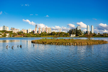 Barigui Park lake and island in Curitiba, Parana, Brazil, showcasing the modern city skyline against a bright blue sky.