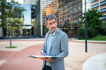 Mature businessman holding clipboard checking documents outdoors