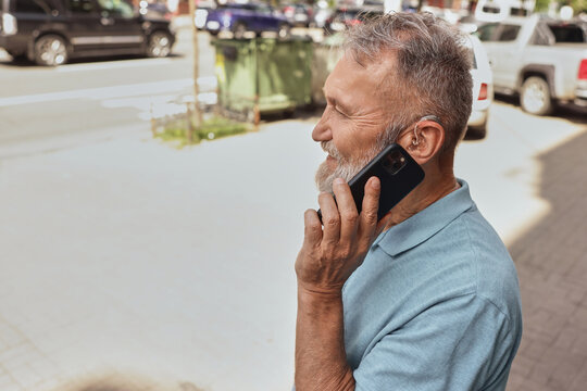 Senior man with a hearing impairment uses a hearing aid in everyday life, talking on phone in urban city outdoor. Hearing solutions