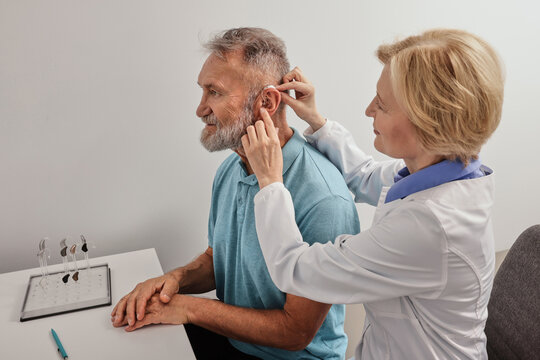 Audiologist putting hearing aid on elderly man's ear at audiology center, close-up, side view. Deafness treatment, hearing solutions