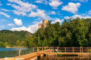 A rustic wooden pier stretches over the water with a view of niedzica castle rising above forested hills. Clear skies and reflections on the lake create a sense of heritage, adventure, and timeless la