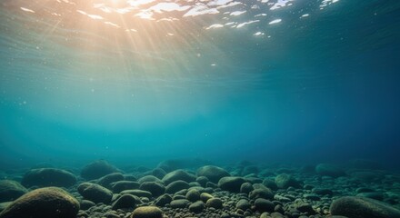 Sunlight streams through clear turquoise water, illuminating smooth rocks and pebbles on the seafloor. Golden rays penetrate the aqua depths, creating a serene underwater environment.