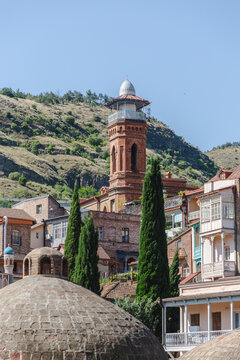 View of the ancient sulfur baths, domes textured like turtle shells, and a minaret reaching for the clear sky, nestled against green hills, Abanotubani, Tbilisi, Georgia.
