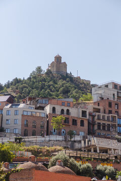 View of ancient fortress perched atop a verdant hill overlooking the city's varied architecture under a clear sky, Abanotubani, Tbilisi, Georgia.