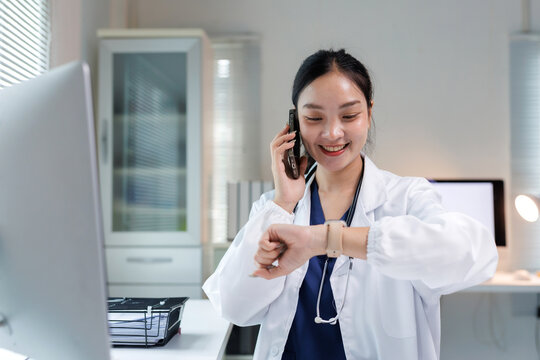 Asian female doctor talking on a mobile phone while checking her smartwatch, managing time and scheduling appointments