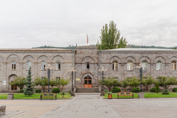 View of aged stone building with arched windows and a small flag fluttering atop, surrounded by verdant trees and benches, Goris, Syunik Province, Armenia.