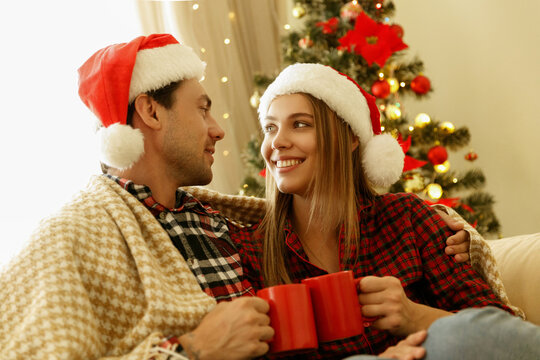 A young couple in Santa hats smiles warmly at each other, wrapped in a cozy blanket and holding red mugs, sitting near a glowing Christmas tree.