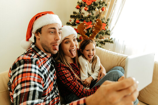 Joyful family making silly faces while taking a selfie with a tablet, dressed in Santa hats and reindeer antlers, sitting by a decorated Christmas tree.