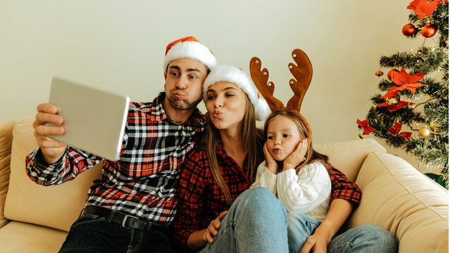 Playful family making funny faces while taking a selfie together, wearing Santa hats and reindeer antlers beside a decorated Christmas tree
