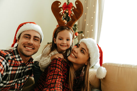 Happy parents and daughter posing together, mother and father in Santa hats, little girl with reindeer antlers, sitting near Christmas tree, sharing love, warmth, and festive family atmosphere.