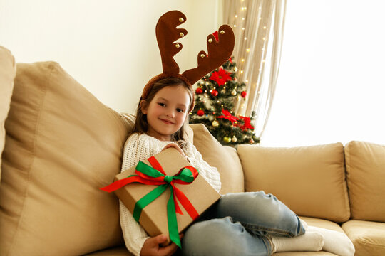 Happy little girl in reindeer antlers holding wrapped Christmas gift with ribbon, sitting on beige sofa near decorated tree with lights. Cozy holiday atmosphere at home.
