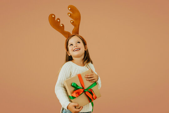 Overjoyed little girl in reindeer antlers holding Christmas gift with ribbon, wearing white sweater and jeans. Festive child portrait on warm background celebrating holiday season.