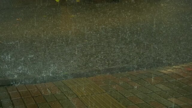 Heavy rain falling on city street asphalt and pavement creating huge puddles. Rainwater cascading over tiled sidewalk and asphalt street during heavy nighttime urban downpour.