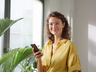 Three-quarter portrait of a European influencer in a minimalist office, she smiles into the camera and holds the smartphone in one hand; yellow shirt