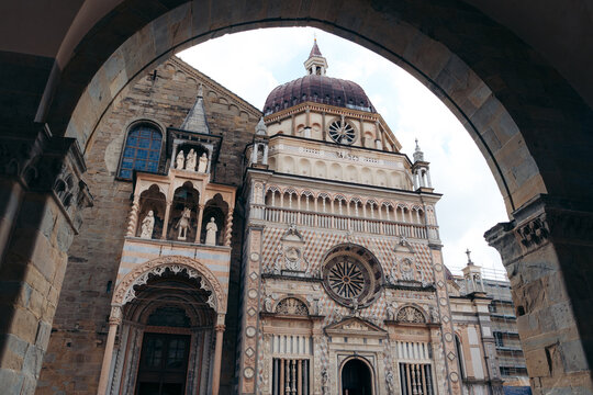 View of sunlit marble facades and intricate stonework frame the Cappella Colleoni and Basilica di Santa Maria Maggiore, under an arched gateway, Bergamo, Lombardy, Italy.
