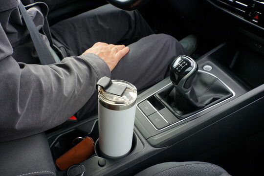 Driver in a car, close-up of manual gear shift lever, white travel mug in cup holder, leather keychain on center console.
