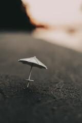 Miniature beach scene with a small umbrella on a sand at sunset. A small, white beach umbrella stands on dark sand, with a blurred sunset in the background, creating a peaceful and serene sunset scene