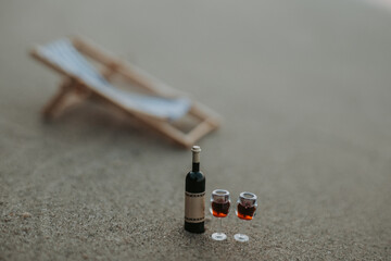 Wine glasses and a bottle of wine on the beach. A miniature wine bottle and two glasses of red wine sit on a sandy beach, with a blurred beach chair in the background.