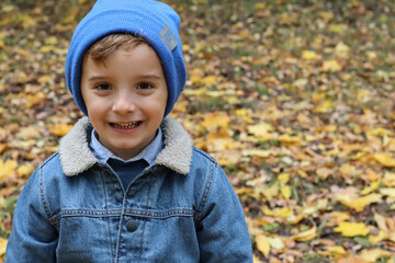 A happy child wearing a blue hat and jacket holds bright yellow autumn leaves in a park filled with vibrant fall colors. 