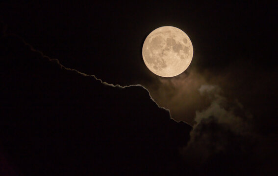 Harvest Super Moon, bathed in golden light, peeks from behind a veil of dark, swirling clouds, creating an ethereal contrast, Lugano Switzerland