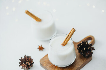 Two glasses of creamy winter drink with cinnamon sticks and pine cones on a board
