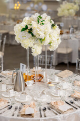 An elegantly served wedding table decorated with tall white flowers in the center creates an elegant and romantic atmosphere of the holiday. A small round table with cutlery and gift boxes for guests