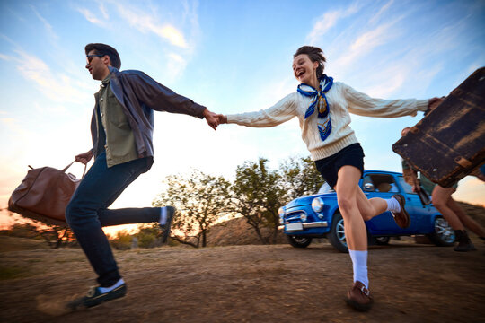Happy couple holding hands and running with suitcases by retro blue car. Concept of love, lifestyle branding, nostalgic travel, freedom and adventurous storytelling. - Powered by Adobe