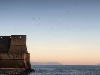 The edge of the Castell dell'Ovo rises from Naples Harbour. Canon are visible through openings in the wall. Gulls circle in the clear sky above the fort. Headlands are in the distance.