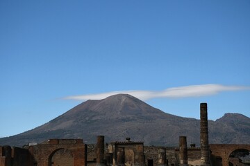 Broken columns and arches from the Forum in Pompeii are seen with Mount Vesuvius in the background. A road is visible winding up the mountain. The sky is blue with a few clouds.
