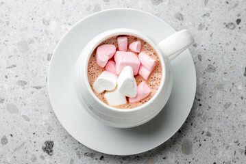 Hot chocolate in a white ceramic cup topped with pink and white heart-shaped marshmallows on a saucer, placed on a gray stone background. Cozy winter drink concept