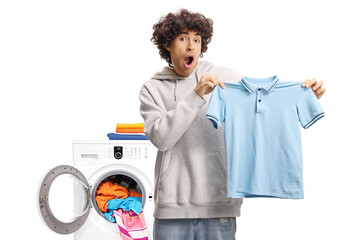 Surprised young man holding a small blue t-shirt in front of a washing machine