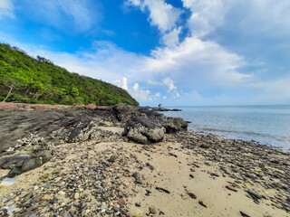 Rocky shoreline with lush green hill and calm ocean under partly cloudy sky, evoking sense of...