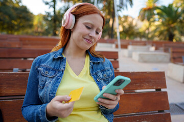 Young woman making an online payment outdoors with a smartphone and credit card, listening to music