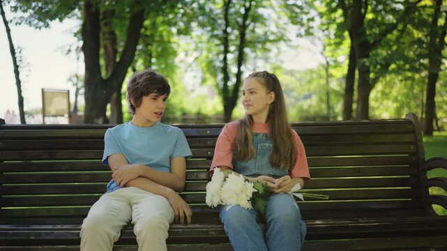 Teen girl with flowers bouquet sitting on bench, talking with boyfriend in park