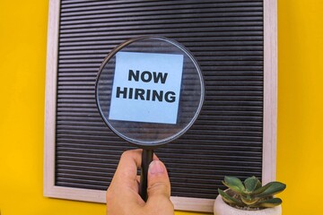 Magnifying Glass Reveals Now Hiring On Letter Board Over Yellow Background