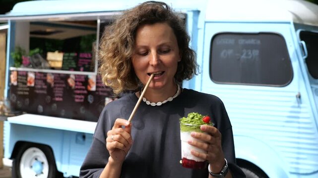 Happy curly haired woman enjoying a healthy layered dessert with matcha and berries from a food truck, playfully biting a biodegradable straw while looking directly at the camera