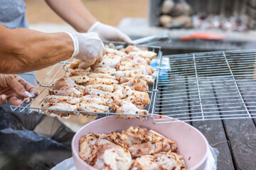 Grilling chicken on a barbecue. A person wearing white gloves is handling marinated chicken pieces on a grill. Outdoor cooking scene in a park.