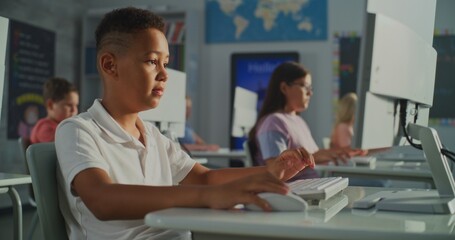 African American Schoolboy in White Polo Shirt is Concentrating on Work at Computer in Modern Classroom. In Background, Children Also Studying at Monitors. Digital Education, Technology. Handheld.