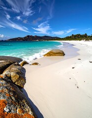 Pristine white sand beach meets turquoise water, large rocks in foreground, mountains in background under a blue sky